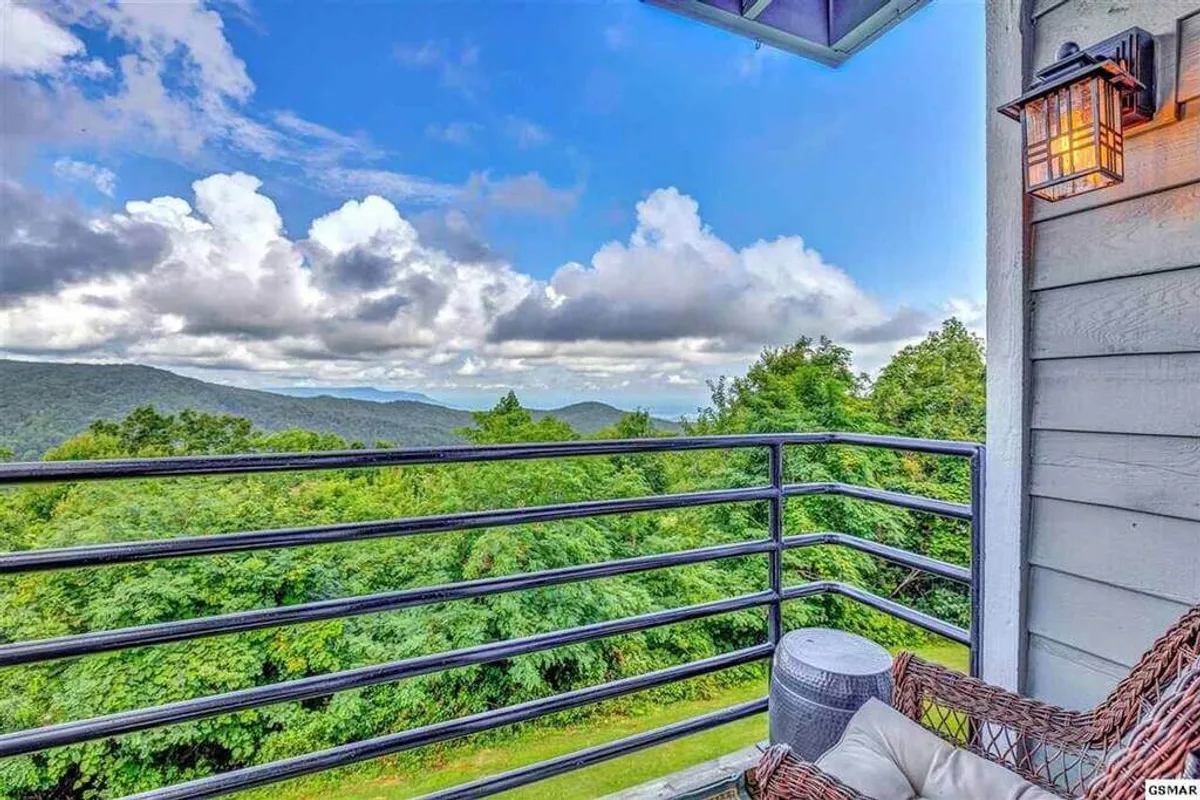 A private balcony furnished with deck furniture overlooks a sweeping mountain view, with a tranquil, cloud-dappled sky above. The serene setting invites relaxation and offers an immersive natural escape.