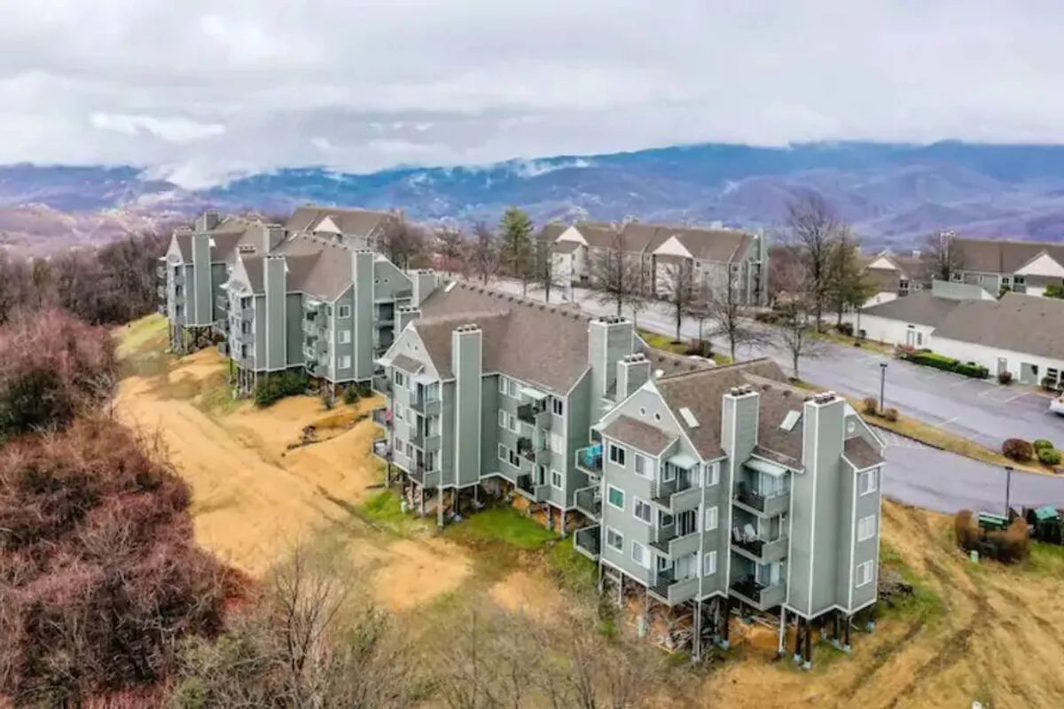A wide-angle view of multi-story condo buildings nestled among landscaped grounds, with a dramatic mountain backdrop rising behind. The scene exudes serenity and a sense of arrival, with open spaces ideal for families to gather outdoors and soak in the natural beauty of the Smokies.