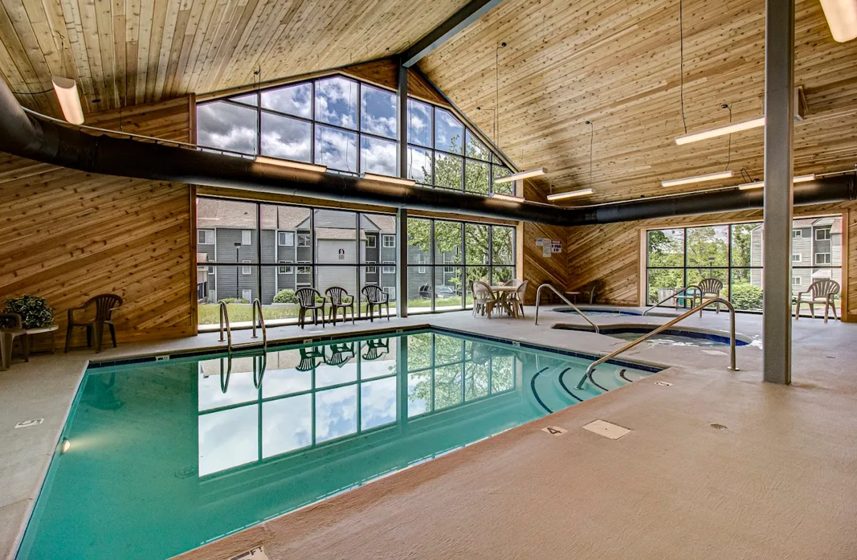 An inviting indoor swimming pool surrounded by large windows and a wooden ceiling, with lounge chairs arranged poolside. Natural light streams in, creating a rejuvenating and peaceful atmosphere.