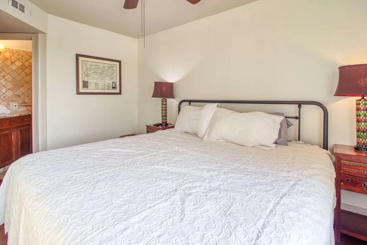 A spacious bedroom with a large bed dressed in white bedding, colorful bedside lamps adding a cheerful touch, and framed artwork on the wall. The room is flooded with natural light, creating a serene environment perfect for resting or reading.