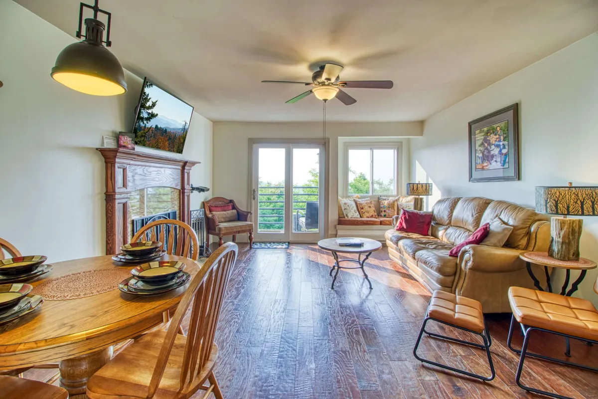 A cozy, open-concept living and dining area featuring a large dining table, a plush sofa, a fireplace, and a viewing area with TV. Natural light filters in, and the nearby balcony offers glimpses of mountain views, creating a warm, inviting hub for gatherings.
